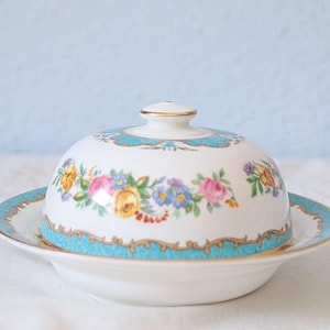 Può includere: Un burriera in porcellana bianca con coperchio a cupola caratterizzato da un colorato motivo floreale con fiori rosa, gialli e blu. La base e il coperchio sono rifiniti con un bordo turchese brillante e finiture dorate sui profili.