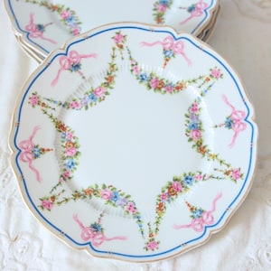Op de afbeelding: Een wit porseleinen bord met een geschulpte rand, voorzien van een blauwe rand en een decoratief patroon van roze linten en kleurrijke bloemenslingers bestaande uit roze, blauwe en gele bloemen in een symmetrisch hangend ontwerp.