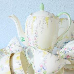 May include: A cream-coloured porcelain tea set featuring a delicate floral pattern with yellow, pink, and blue blossoms. The set includes a tall teapot with a light green handle and lid finial, several matching teacups, and saucers with gold trim.