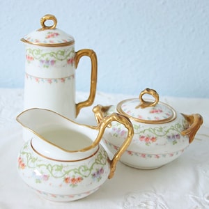 May include: A porcelain tea set consisting of a lidded pitcher, a creamer, and a lidded sugar bowl. Each piece is white with a delicate floral pattern in pink, blue, and green, accented by ornate gold handles and matching gold finials.