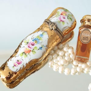 Op de afbeelding: Een goudomrand porseleinen scharnierdoosje met handgeschilderde roze, blauwe en oranje bloemmotieven, vergezeld door een klein glazen parfumflesje met het label Calèche Hermès en een streng witte parels met een glanzende afwerking.