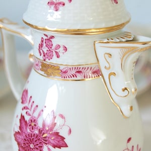 May include: White porcelain coffee pot with a floral design in shades of pink and gold. The pot has a textured lid and handle, with a decorative spout. A classic piece of tableware.