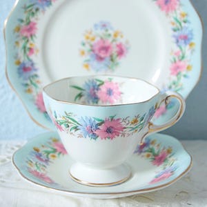 May include: A vintage tea set featuring a teacup, saucer, and plate. The set has a light blue border with pink and blue floral designs and gold trim. The teacup is white with a pedestal base.