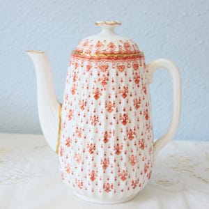 Op de afbeelding: Witte keramische koffiepot met een deksel, versierd met een herhalend oranje fleur-de-lis patroon. De pot heeft een gebogen handvat, een lange tuit en gouden randen. De achtergrond is lichtblauw.