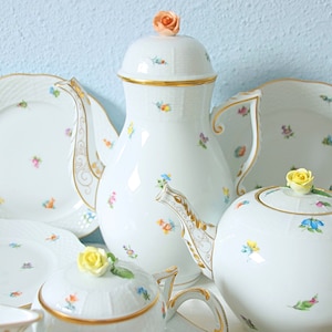 Op de afbeelding: Een wit porseleinen theeservies met delicate bloempatronen en gouden randen. De set bevat een theepot, een suikerpot en borden. Elk stuk is versierd met kleine, kleurrijke bloemmotieven.