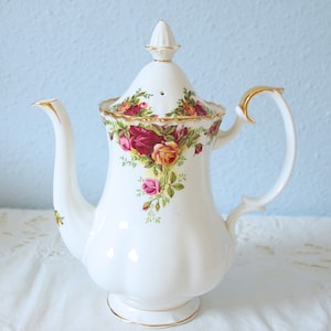 Op de afbeelding: Een witte porseleinen theepot met een geschulpte voet en een bloemmotief van rode, gele en roze rozen met groene bladeren. De theepot is voorzien van een bijpassend deksel met een puntige knop, een gebogen handvat en een tuit met gouden randen.