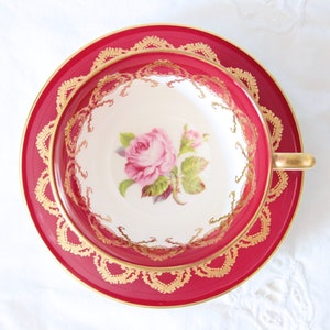 Può includere: Un set di tazza da tè e piattino in porcellana con un bordo rosso cremisi intenso e intricati filigrani dorati. Il centro della tazza bianca presenta un'illustrazione dettagliata di una rosa rosa con foglie verdi e un piccolo bocciolo.