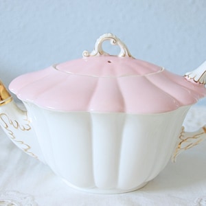 May include: A white ceramic teapot with a scalloped body and a matching pale pink lid. The teapot features a gold-toned spout, a decorative handle with gold accents, and delicate gold filigree detailing along the base of the spout and handle.