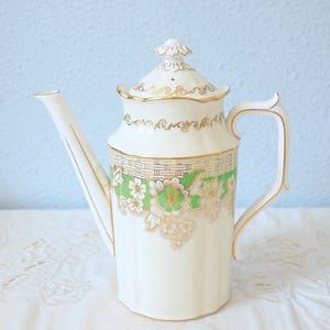 Op de afbeelding: Een crèmekleurige porseleinen theepot met een groene band, witte bloemmotieven en gouden randen. De theepot heeft een hoge, slanke tuit, een gebogen handvat en een bijpassend deksel met een decoratieve gouden knop als bekroning.