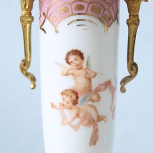 Könnte beinhalten: Eine weiße Porzellanvase mit zwei goldfarbenen Metallgriffen an den Seiten. Der obere Rand ist mit einem rosa-goldenen Muster verziert. In der Mitte der Vase befindet sich eine gemalte Abbildung von zwei geflügelten Putten mit rosa Bändern.