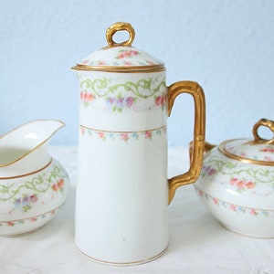 Op de afbeelding: Een wit porseleinen koffieservies met gouden accenten. De set bestaat uit een hoge koffiepot met deksel, een melkkan en een suikerpot. Elk stuk is versierd met een bloemmotief in pastelkleuren en groene slingers.