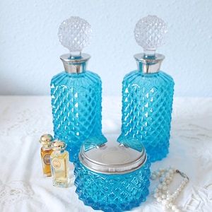 May include: Two tall, bright turquoise glass decanters with diamond-patterned bodies and clear, spherical, faceted stoppers, accompanied by a matching turquoise glass jar with a silver lid and two small amber-colored glass perfume bottles.