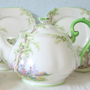 Op de afbeelding: Een witte porseleinen theepot met een geschulpte rand, een felgroen handvat en een geschilderd landschap met bomen en kleurrijke bloemen. De theepot heeft een bijpassend deksel met een groene knop en een gebogen tuit.