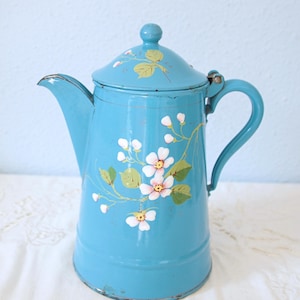 Op de afbeelding: Een felblauwe geëmailleerde metalen koffiepot met een handgeschilderd bloemmotief met witte bloemblaadjes, gele harten en groene bladeren. De pot heeft een bijpassend deksel met een kleine knop, een gebogen handvat en een tuit met zichtbare slijtage.