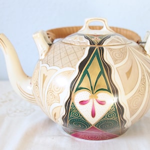 Può includere: Teiera in ceramica color crema con un design decorativo in oro, verde e marrone. La teiera presenta un beccuccio curvo, un manico intrecciato e un coperchio con una piccola maniglia. Il design include motivi geometrici e floreali.