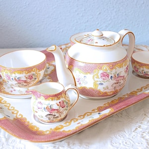 Op de afbeelding: Een vintage theeservies met een roze en gouden bloemenontwerp. De set bevat een theepot, een theekopje, een schotel, een melkkan en een kleine kom, allemaal gerangschikt op een bijpassend dienblad. De theeset is gemaakt van porselein.