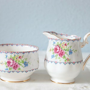 Op de afbeelding: Een witte porseleinen melk- en suikerpotset. De set heeft een kruissteek bloemenontwerp in roze, blauw en groen. Gouden randen accentueren de randen en handvatten. De suikerpot is 10 cm breed.