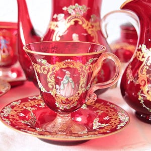 Op de afbeelding: Een theeservies van robijnrood glas met gouden filigraanranden en handgeschilderde scènes van figuren in historische kleding. De set bevat een theepot, een melkkan en meerdere theekopjes met bijpassende schoteltjes, allemaal voorzien van gouden randen.