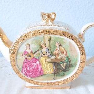 Op de afbeelding: Een witte porseleinen theepot met een centraal ovaal medaillon met een kleurrijke illustratie van drie personen in historische kleding, van wie er één luit speelt. De theepot is afgewerkt met gouden accenten, een gouden handvat, tuit en strikvormige dekselknop.