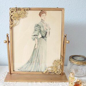 May include: A rectangular brass swivel picture frame featuring ornate butterfly-shaped corner accents. The frame holds a vintage-style illustration of a person wearing a long, pale blue dress with ruffled sleeves and a high collar, standing in profile.