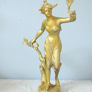 Op de afbeelding: Goudkleurig beeld van een vrouw in een vloeiende jurk, die een tak met bladeren vasthoudt. Het beeld heeft een klassiek ontwerp en is op een voetstuk gemonteerd. De achtergrond is een lichtblauwe muur.
