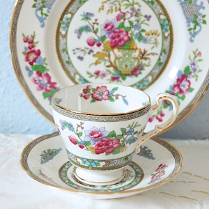 Könnte beinhalten: Ein Vintage-Teeservice mit einer Tasse, Untertasse und Teller. Die weißen Porzellanstücke sind mit einem Blumenmuster in Rosa, Rot, Blau und Grün verziert und mit Gold verziert. Das Set ist für Teepartys gedacht.