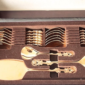 May include: A gold-toned cutlery set in a brown presentation box. The set includes forks, spoons, and a cake server, each featuring a detailed floral motif on the handle. The utensils are arranged in a moulded brown velvet-lined tray.