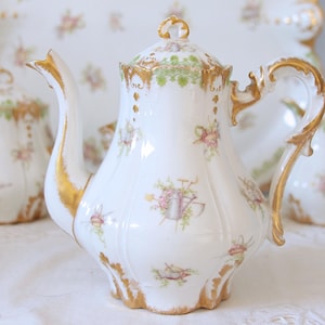 Op de afbeelding: Witte porseleinen theepot met gouden rand en bloemen- en tuingereedschapmotieven. De theepot heeft een gebogen tuit en handvat, en een decoratief deksel. Het ontwerp bevat roze bloemen en groene bladeren.