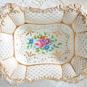 Op de afbeelding: Een sierlijke, rechthoekige witte porseleinen schaal met gouden randen en bloemaccenten. De schaal heeft een handgeschilderd bloemstuk in het midden, met roze, blauwe en paarse bloemen. De randen hebben een gedetailleerd, gebeeldhouwd ontwerp.