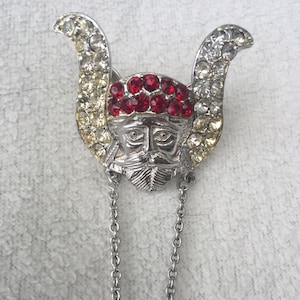 May include: A silver-toned brooch with a face and red and clear rhinestones. The brooch has a dangling chain with a smaller version of the brooch at the end.