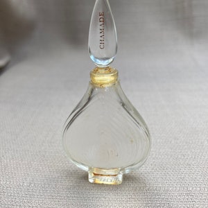 May include: A clear glass perfume bottle with a textured surface and a pointed stopper. The stopper is inscribed with the word "CHAMADE" in gold lettering.