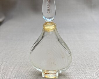 Può includere: Un flacone di profumo in vetro trasparente con una superficie testurizzata e un tappo a punta. Il tappo è inciso con la parola "CHAMADE" in lettere dorate.