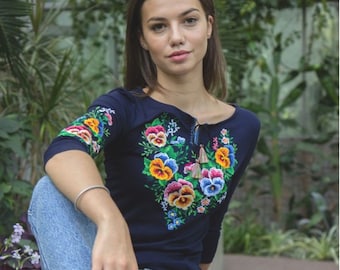 May include: A woman wearing a navy blue long-sleeved shirt with embroidered flowers and a pair of blue jeans. She is sitting on a stone wall and wearing black and white sneakers.