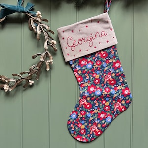 May include: A Christmas stocking featuring a navy blue fabric body with a dense floral pattern in red, blue, and yellow. The cuff is made of light beige fabric with the name Georgina embroidered in red thread, accented by small red star motifs.
