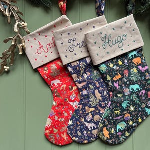 May include: Three Christmas stockings with beige cuffs featuring embroidered names and small star accents. The stockings have colourful fabric patterns depicting various animals, trees, and gifts on red, dark blue, and black backgrounds respectively.