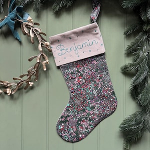 May include: A Christmas stocking featuring a dark fabric body with a dense, colourful pattern of small stars and circular motifs. The cuff is a light mauve fabric with the name Benjamin embroidered in teal thread, and a matching fabric hanging loop.