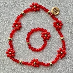 Op de afbeelding: Een set van twee rode kralenarmbanden met bloembedels. De grotere armband heeft een gouden label met de tekst "Neon Barbie".