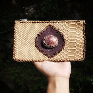 Pode incluir: Uma pequena bolsa de couro bege com fecho de correr. A bolsa tem uma aba de couro castanho com um padrão de costura decorativo e uma gema rosa no centro.