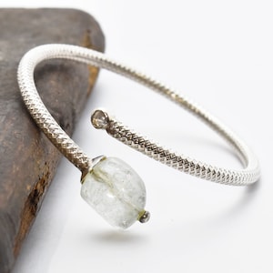 Può includere: Un braccialetto rigido in argento con una pietra preziosa trasparente e sfaccettata. Il braccialetto è attorcigliato e presenta un design testurizzato.