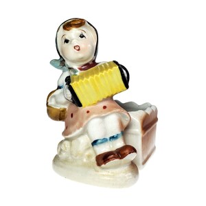 May include: A ceramic figurine of a seated individual wearing a headscarf and a polka-dot dress, playing a bright yellow accordion. The figure is positioned next to a brown rectangular container, all finished in a glossy, hand-painted glaze.