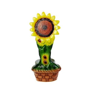 May include: A ceramic napkin ring featuring a three-dimensional sunflower with bright yellow petals and a textured brown center. The stem is green with two small sunflower buds, and the base is shaped like a woven brown basket with a glossy finish.