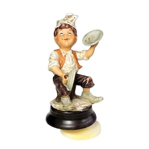 May include: A porcelain figurine of a seated child wearing a white shirt, orange vest, and brown trousers, topped with a white cap. The figure holds two small, light-colored circular plates in their hands, positioned on a dark circular base.