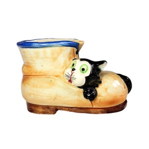 May include: A ceramic planter shaped like a beige shoe with a dark brown sole and a blue rim. A black and white cat with bright green eyes and a pink nose is sculpted onto the side of the shoe, appearing to peek out from the opening of the footwear.