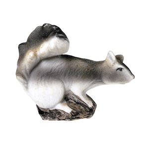 May include: A small ceramic squirrel figurine featuring a white body with shaded gray and black accents on the tail, back, and head. The squirrel is depicted in a seated position on a textured, dark brown base that mimics a natural rock surface.