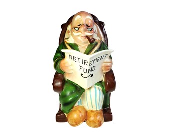 May include: A ceramic figurine of an elderly person with a mustache and glasses, wearing a green robe and striped pants, sitting in a brown rocking chair. The figure holds an open white newspaper with the text RETIREMENT FUND and smokes a pipe.