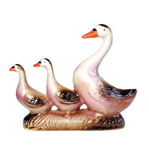 May include: A ceramic figurine featuring three geese of varying sizes standing together on a textured brown base. The geese have white heads, orange beaks, and bodies with a glossy finish in shades of light pink, tan, and dark brown accents.