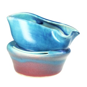 May include: A two-piece ceramic gravy boat and base set featuring a glossy blue glaze. The top piece is an elongated, curved vessel with a pouring spout, while the bottom piece is a wide, bowl-shaped stand with a reddish-brown base transition.