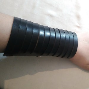 May include: Black leather wrist cuff with multiple horizontal straps. The cuff is secured with small silver-colored rivets. The image shows the cuff worn on a person's wrist, with the hand and fingers visible.