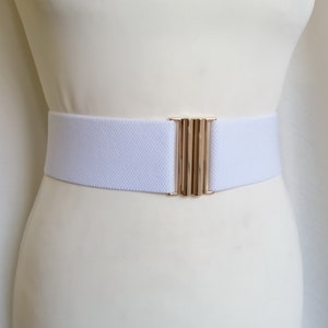 May include: A wide white elastic belt featuring a polished gold-tone metal clasp closure. The belt is constructed from a textured fabric material and is designed to be worn around the waist, providing a secure fit with its flexible, stretchy band.