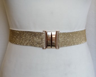 May include: A gold-coloured belt with a metallic mesh design and a silver-coloured rectangular clasp. The belt is displayed on a white mannequin. The clasp has three vertical bars.